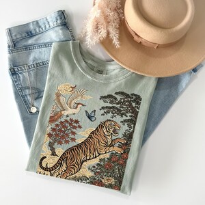 May include: A sage green t-shirt featuring a graphic print of a tiger, a bird, a butterfly, trees, and clouds. The shirt is paired with light blue denim jeans, a tan wide-brimmed hat, and a silver-toned necklace with a small circular pendant.
