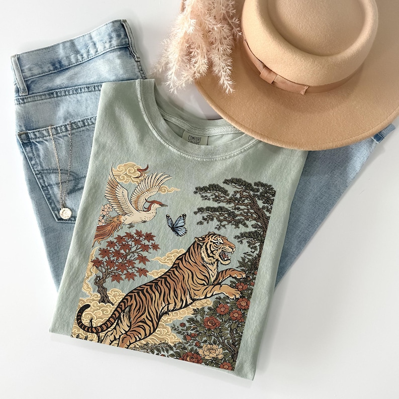May include: A sage green t-shirt featuring a graphic print of a tiger, a bird, a butterfly, trees, and clouds. The shirt is paired with light blue denim jeans, a tan wide-brimmed hat, and a silver-toned necklace with a small circular pendant.