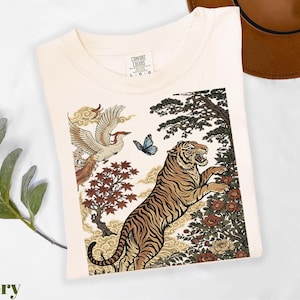 May include: An ivory t-shirt featuring a graphic print of a tiger, a flying bird, a blue butterfly, and floral elements. The shirt has a crew neckline and a Comfort Colors label at the inner neck with the size L-G-G clearly visible on the tag.