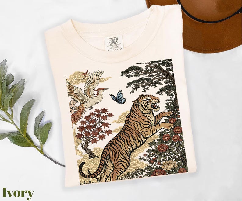 May include: An ivory t-shirt featuring a graphic print of a tiger, a flying bird, a blue butterfly, and floral elements. The shirt has a crew neckline and a Comfort Colors label at the inner neck with the size L-G-G clearly visible on the tag.