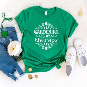 Op de afbeelding: Een groen T-shirt met witte tekst die "Gardening is my therapy" zegt en een afbeelding van tuingereedschap en bladeren.