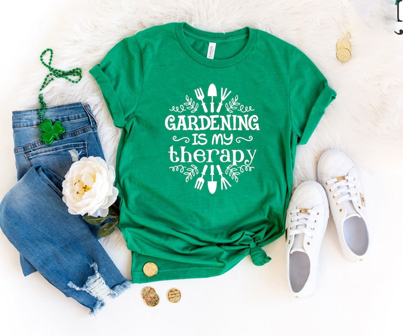 Op de afbeelding: Een groen T-shirt met witte tekst die "Gardening is my therapy" zegt en een afbeelding van tuingereedschap en bladeren.