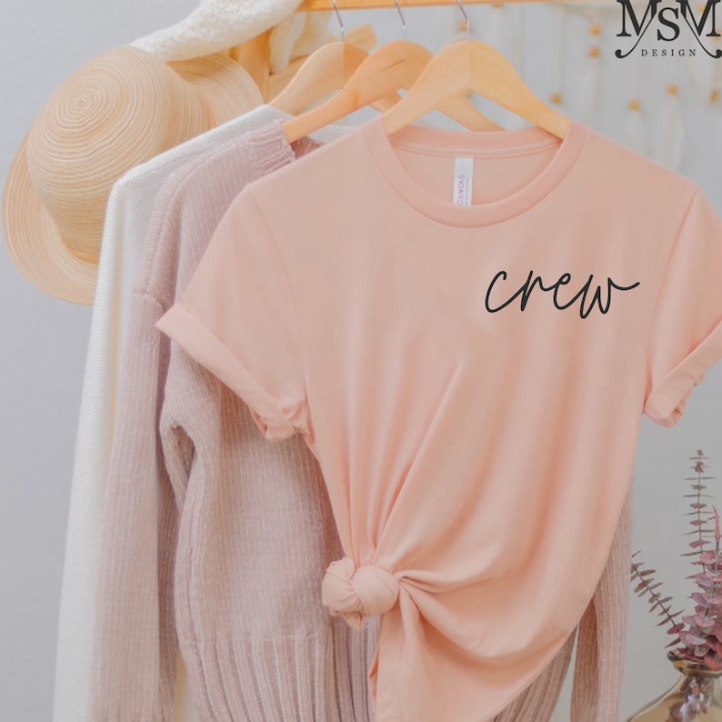 K&ouml;nnte beinhalten: Ein pfirsichfarbenes T-Shirt mit dem Wort "crew" in schwarzer Schrift. Das T-Shirt ist am unteren Ende geknotet und h&auml;ngt an einem Holzb&uuml;gel. Weitere Kleidungsst&uuml;cke, darunter ein wei&szlig;er Pullover und ein rosa Pullover, h&auml;ngen ebenfalls an B&uuml;geln.