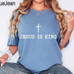 K&ouml;nnte beinhalten: Hellblaues T-Shirt mit dem wei&szlig;en Text "JESUS IS KING" und einem wei&szlig;en Kreuz. Das Shirt hat kurze &Auml;rmel und einen Rundhalsausschnitt. Der Markenname "BlueJean" befindet sich oben links.
