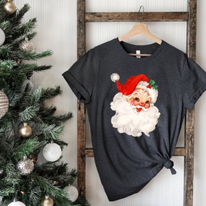 May include: Dark gray t-shirt featuring a vintage Santa Claus graphic. The Santa Claus illustration has a red hat, white beard, and rosy cheeks. The shirt is hanging on a wooden hanger against a white background.