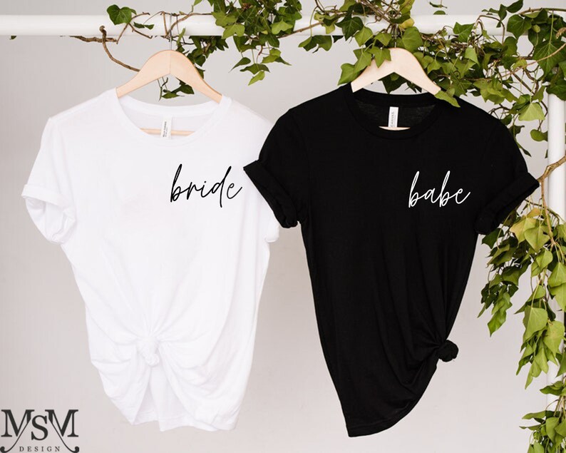 K&ouml;nnte beinhalten: Zwei T-Shirts auf Holzb&uuml;geln. Eines ist wei&szlig; mit dem Wort "bride" in schwarzer Schrift. Das andere ist schwarz mit dem Wort "babe" in wei&szlig;er Schrift. Gr&uuml;nes Laub befindet sich im Hintergrund.