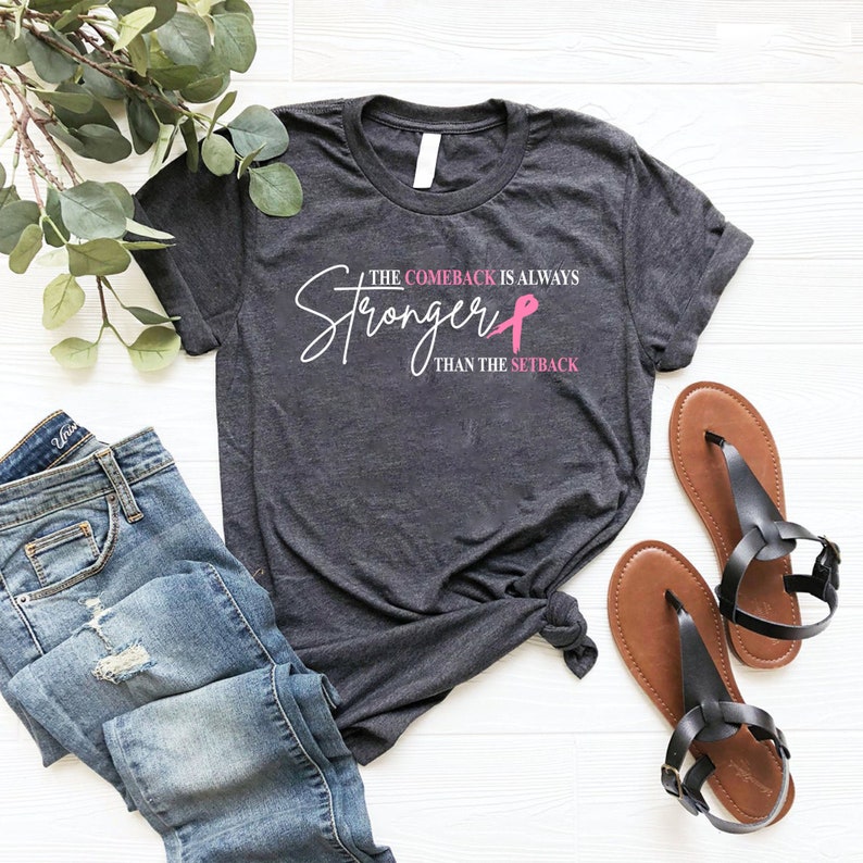 May include: Dark gray t-shirt with the text "The Comeback Is Always Stronger Than The Setback" in white and pink. The shirt is paired with blue jeans and brown sandals. A pink ribbon graphic is included.