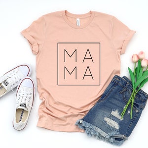 May include: A peach-colored t-shirt with the word "MAMA" in a black square. The shirt is paired with white sneakers, denim shorts, and a bouquet of pink tulips. The shirt is a casual style.