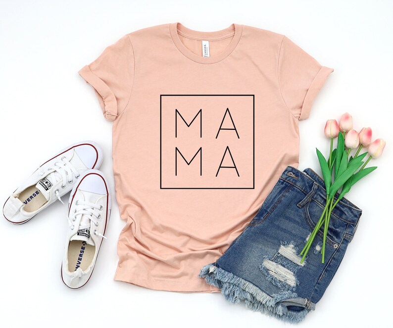 May include: A peach-colored t-shirt with the word "MAMA" in a black square. The shirt is paired with white sneakers, denim shorts, and a bouquet of pink tulips. The shirt is a casual style.