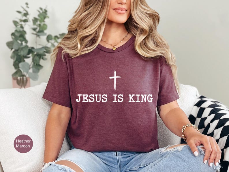 K&ouml;nnte beinhalten: Burgunderfarbenes T-Shirt mit dem wei&szlig;en Text "JESUS IS KING" und einem wei&szlig;en Kreuz. Das T-Shirt wird mit blauen Jeans kombiniert. Es ist ein Freizeitkleidungsst&uuml;ck. Das T-Shirt ist aus weichem Material.