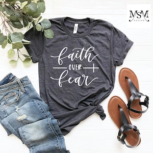May include: A dark gray t-shirt with the text "faith over fear" in white lettering. The shirt is laid out on a white surface with a pair of brown sandals.