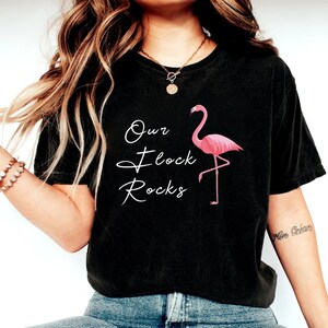 K&ouml;nnte beinhalten: Schwarzes T-Shirt mit dem Text "Our Flock Rocks" in wei&szlig;er Schrift und einer rosa Flamingo-Grafik. Der Flamingo steht auf einem Bein. Das T-Shirt hat einen l&auml;ssigen Stil.