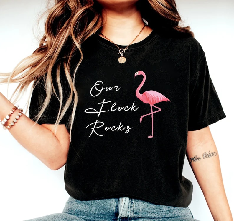 K&ouml;nnte beinhalten: Schwarzes T-Shirt mit dem Text "Our Flock Rocks" in wei&szlig;er Schrift und einer rosa Flamingo-Grafik. Der Flamingo steht auf einem Bein. Das T-Shirt hat einen l&auml;ssigen Stil.