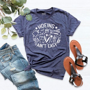 May include: A navy blue t-shirt with a white graphic that says "HOEING AIN'T EASY" and features a variety of gardening tools and plants.