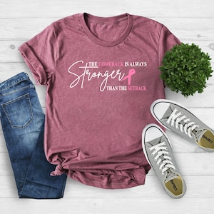 May include: A maroon t-shirt with the text "The Comeback Is Always Stronger Than The Setback" in white script and a pink ribbon graphic. The shirt is paired with blue jeans and gray sneakers.