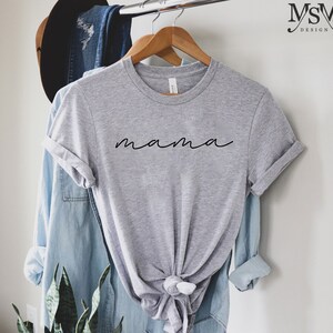 Puede incluir: Una camiseta gris jaspeada con la palabra "mama" en fuente negra. La camiseta est&aacute; colgada en una percha de madera, con una camisa vaquera azul claro y un sombrero marr&oacute;n en el fondo.