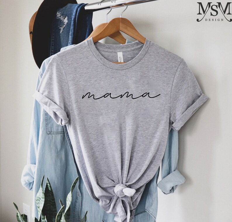Puede incluir: Una camiseta gris jaspeada con la palabra "mama" en fuente negra. La camiseta est&aacute; colgada en una percha de madera, con una camisa vaquera azul claro y un sombrero marr&oacute;n en el fondo.