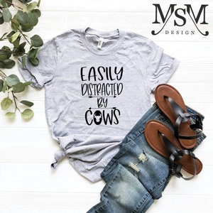 May include: A light gray t-shirt with the text "EASILY DISTRACTED BY COWS" in black. The shirt is paired with blue denim jeans and brown sandals with black straps. The image is on a white surface.