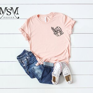 Peut inclure: Un t-shirt rose clair avec un motif de champignon noir, associ&eacute; &agrave; un jean bleu et des baskets blanches. Le t-shirt est nou&eacute; &agrave; la taille. Des baskets blanches en toile compl&egrave;tent l'ensemble d&eacute;contract&eacute;.