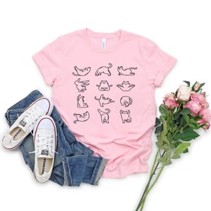 Op de afbeelding: Een roze T-shirt met een afbeelding van katten die yoga-poses aannemen. Het shirt ligt op een wit oppervlak met een paar witte sneakers en een bos roze rozen.