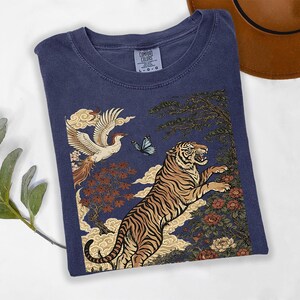 May include: A navy blue short-sleeve t-shirt featuring a graphic print of a leaping tiger, a flying phoenix, a blue butterfly, stylized clouds, and floral elements. The shirt has a crew neckline and a Comfort Colors brand label at the inner collar.