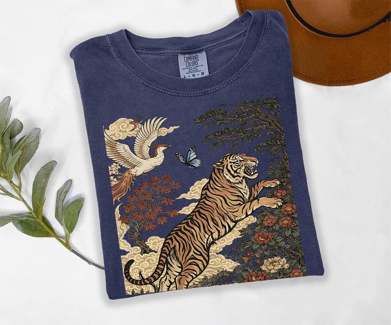 May include: A navy blue short-sleeve t-shirt featuring a graphic print of a leaping tiger, a flying phoenix, a blue butterfly, stylized clouds, and floral elements. The shirt has a crew neckline and a Comfort Colors brand label at the inner collar.
