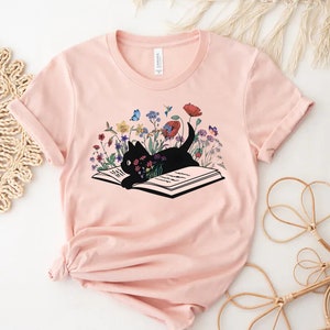 Pu&ograve; includere: Una maglietta rosa con un gatto nero rannicchiato su un libro aperto. Il libro &egrave; circondato da fiori colorati e farfalle.