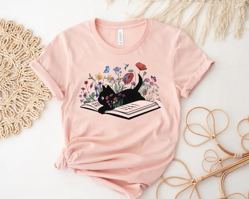 Pu&ograve; includere: Una maglietta rosa con un gatto nero rannicchiato su un libro aperto. Il libro &egrave; circondato da fiori colorati e farfalle.