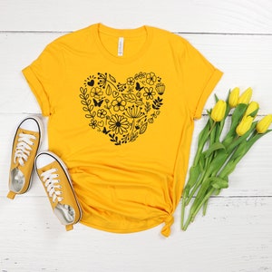 May include: A yellow t-shirt with a black floral heart design. The shirt is laid out on a white wooden surface with a pair of yellow sneakers and a bouquet of yellow tulips.