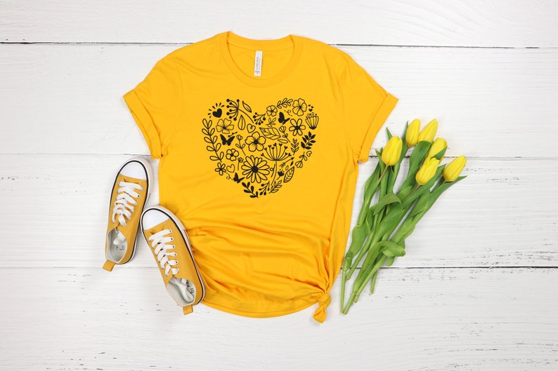 May include: A yellow t-shirt with a black floral heart design. The shirt is laid out on a white wooden surface with a pair of yellow sneakers and a bouquet of yellow tulips.