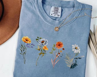 May include: A blue jean Comfort Colors t-shirt with a floral design. The shirt features a watercolor print of various flowers in shades of orange, yellow, purple, and white. A gold necklace with a pendant is draped over the shirt.