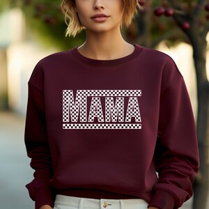 Puede incluir: Una sudadera granate con la palabra "MAMA" en letras blancas a cuadros. Las letras est&aacute;n delineadas en blanco. La sudadera tiene cuello redondo.