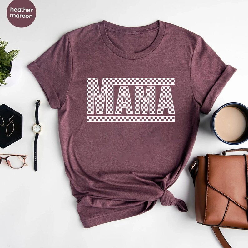 Puede incluir: Camiseta color granate jaspeado con la palabra "MAMA" en un dise&ntilde;o a cuadros blancos. La camiseta tiene cuello redondo y mangas cortas. La tela es suave.