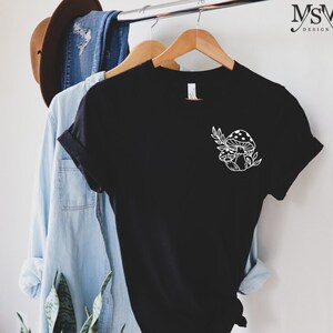 Peut inclure: T-shirt noir avec un motif de champignon et de feuilles blanches. Le t-shirt est suspendu &agrave; un cintre en bois. Une chemise en jean bleu clair et un chapeau marron sont &eacute;galement visibles.