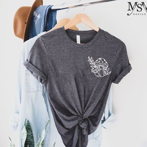 Peut inclure: T-shirt gris fonc&eacute; avec un motif de champignon et de feuilles blanches. Le t-shirt est suspendu &agrave; un cintre en bois avec d'autres v&ecirc;tements. Le texte "MSM DESIGN" est dans le coin sup&eacute;rieur droit.