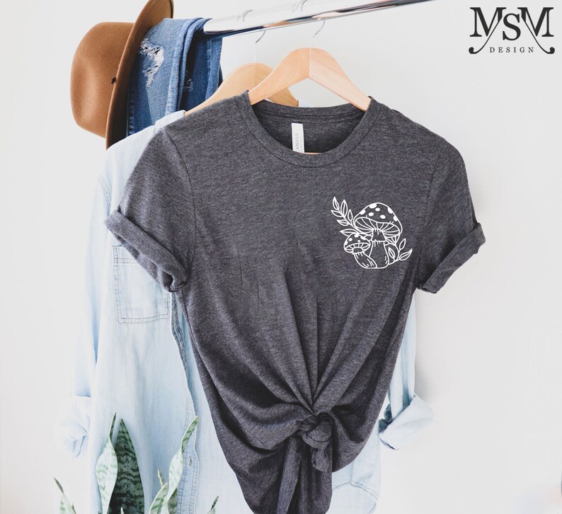 Peut inclure: T-shirt gris fonc&eacute; avec un motif de champignon et de feuilles blanches. Le t-shirt est suspendu &agrave; un cintre en bois avec d'autres v&ecirc;tements. Le texte "MSM DESIGN" est dans le coin sup&eacute;rieur droit.