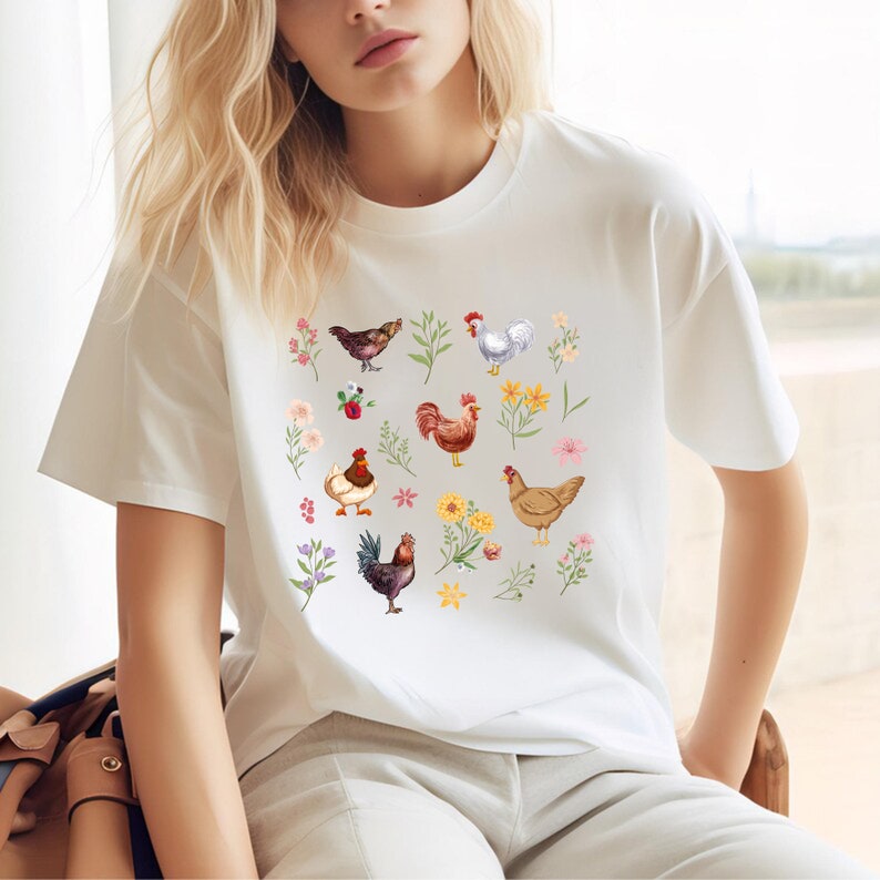 May include: White t-shirt with a colorful pattern of chickens and flowers. The chickens are brown, white, and red with yellow beaks. The flowers are pink, yellow, and blue.