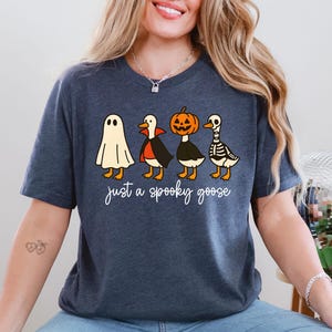 Puede incluir: Camiseta azul marino con un dise&ntilde;o de Halloween. El gr&aacute;fico muestra cuatro gansos disfrazados: un fantasma, un vampiro, una calabaza y un esqueleto. El texto "just a spooky goose" est&aacute; escrito en cursiva debajo.