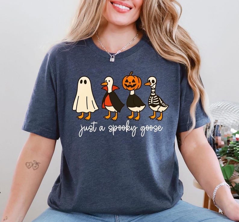 Puede incluir: Camiseta azul marino con un dise&ntilde;o de Halloween. El gr&aacute;fico muestra cuatro gansos disfrazados: un fantasma, un vampiro, una calabaza y un esqueleto. El texto "just a spooky goose" est&aacute; escrito en cursiva debajo.