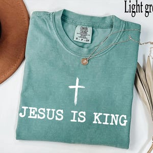 K&ouml;nnte beinhalten: Hellgr&uuml;nes T-Shirt mit dem wei&szlig;en Text "JESUS IS KING" und einem wei&szlig;en Kreuz. Eine silberne Halskette mit Anh&auml;nger liegt auf dem T-Shirt. Das T-Shirt ist aus weichem Material.