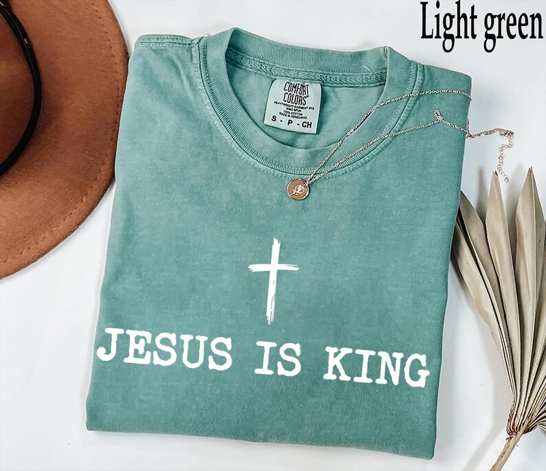 K&ouml;nnte beinhalten: Hellgr&uuml;nes T-Shirt mit dem wei&szlig;en Text "JESUS IS KING" und einem wei&szlig;en Kreuz. Eine silberne Halskette mit Anh&auml;nger liegt auf dem T-Shirt. Das T-Shirt ist aus weichem Material.
