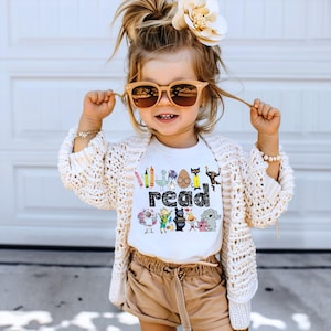 May include: A white t-shirt featuring a colorful graphic print of various cartoon characters surrounding the word read in black text. The shirt is paired with a cream-colored open-knit cardigan, tan shorts, light-colored sunglasses, and a hair accessory.