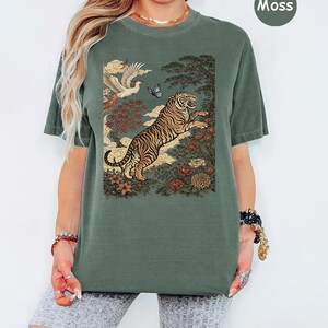 May include: A moss green short-sleeve t-shirt featuring a graphic print of a leaping tiger, a flying bird, a butterfly, and various flowers and trees. The design is set against a muted background, and the shirt has a crew neckline.