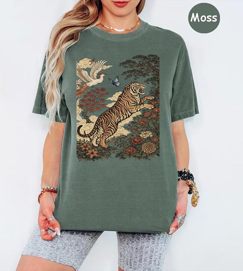May include: A moss green short-sleeve t-shirt featuring a graphic print of a leaping tiger, a flying bird, a butterfly, and various flowers and trees. The design is set against a muted background, and the shirt has a crew neckline.
