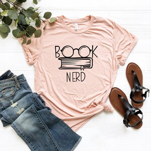May include: A light pink t-shirt with the words "BOOK NERD" in black, featuring glasses over a book. The shirt is paired with blue jeans and black sandals, with a sprig of greenery in the background.