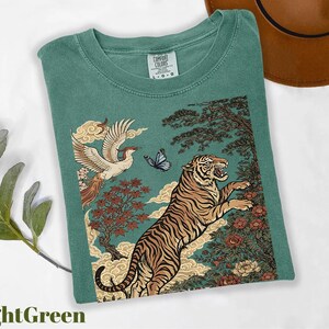 May include: A light green t-shirt featuring a graphic print of a tiger leaping toward a flying bird and a butterfly. The design includes stylized trees, flowers, and clouds in shades of orange, brown, and blue. The shirt has a Comfort Colors label.