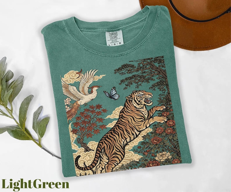 May include: A light green t-shirt featuring a graphic print of a tiger leaping toward a flying bird and a butterfly. The design includes stylized trees, flowers, and clouds in shades of orange, brown, and blue. The shirt has a Comfort Colors label.