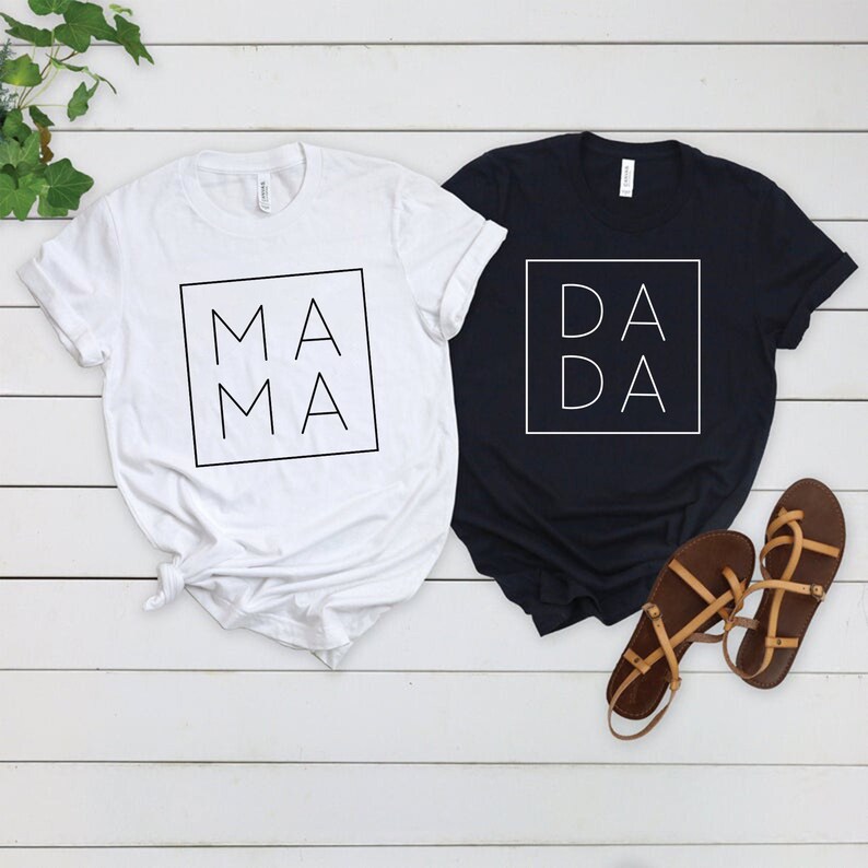May include: Two white t-shirts with black text. One shirt says "MA MA" in a square and the other says "DA DA" in a square.