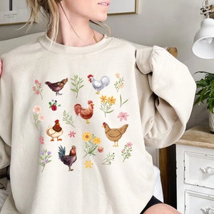 May include: A cream-colored sweatshirt with a graphic print of chickens and flowers. The chickens are brown, white, and red. The flowers are pink, yellow, and purple.