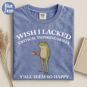 May include: A blue jean colored t-shirt featuring a graphic of a green frog holding a small orange mug. The text above the frog reads WISH I LACKED CRITICAL THINKING SKILLS and the text below the frog reads Y'ALL SEEM SO HAPPY in white font.